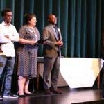 A small group of people stand together on a stage with one student at a microphone and Dr. Jones and School Board President Rankin watching.